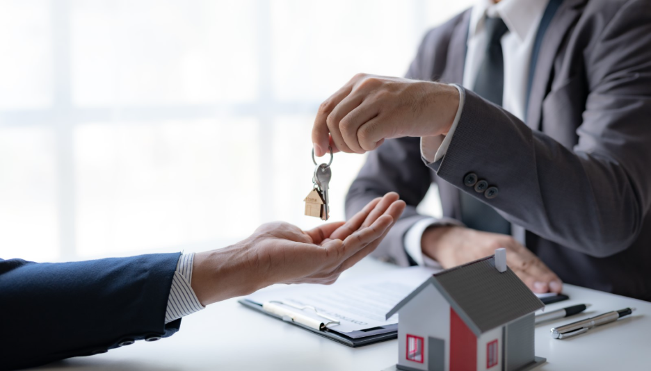 Business professionals exchanging property keys at a desk with a model house and real estate documents, representing a commercial property handover or tenant transition.